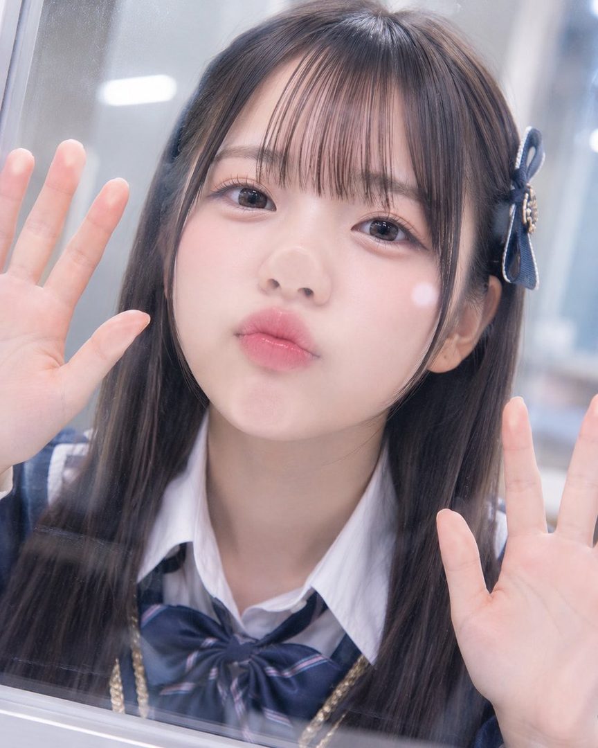 A young girl in a school uniform presses her hands and face against a glass surface, making a pouting expression.