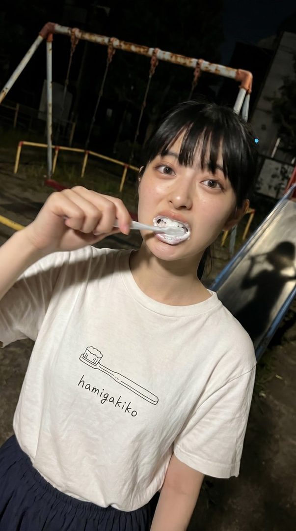 A young woman with black hair and bangs, wearing a white t-shirt with a toothbrush graphic, brushes her teeth at night in an outdoor playground.