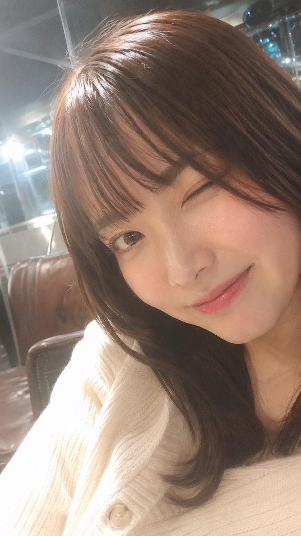 A young woman with brown hair winks at the camera, wearing a cream-colored sweater indoors.