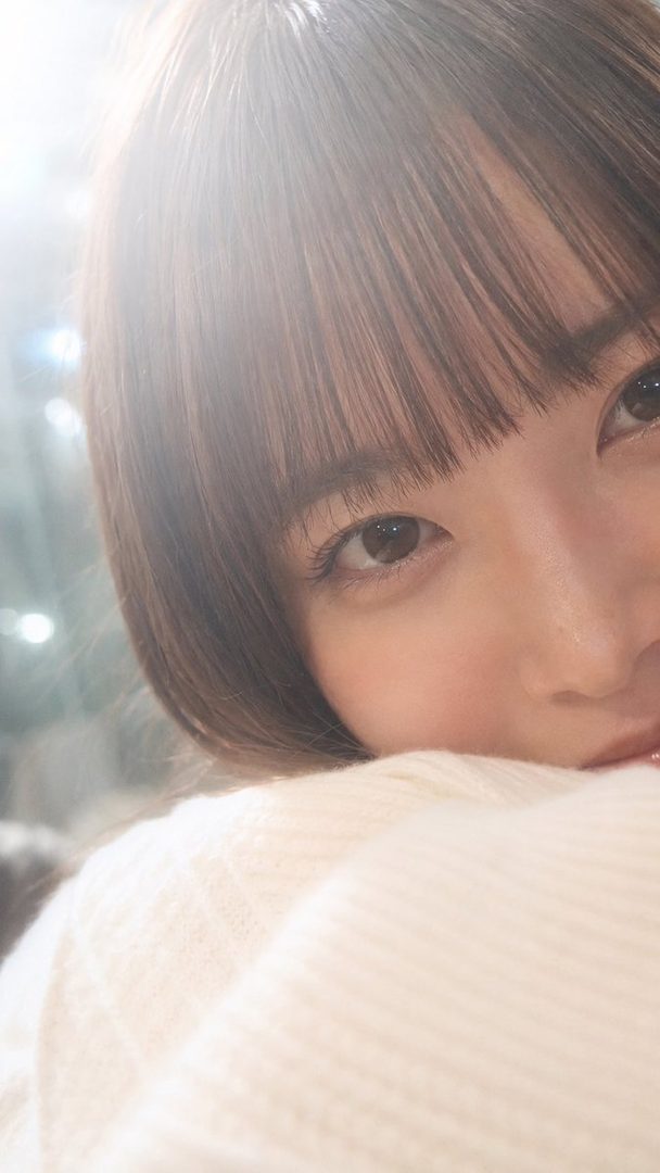 Close-up portrait of a young East Asian woman with brown hair and bangs, wearing a white sweater, looking towards the camera with a soft expression.