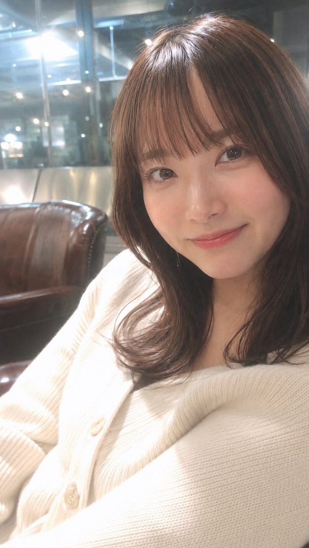 A young East Asian woman with brown hair and bangs, wearing a cream-colored knit cardigan, smiles gently at the camera in an indoor setting with a leather chair visible.