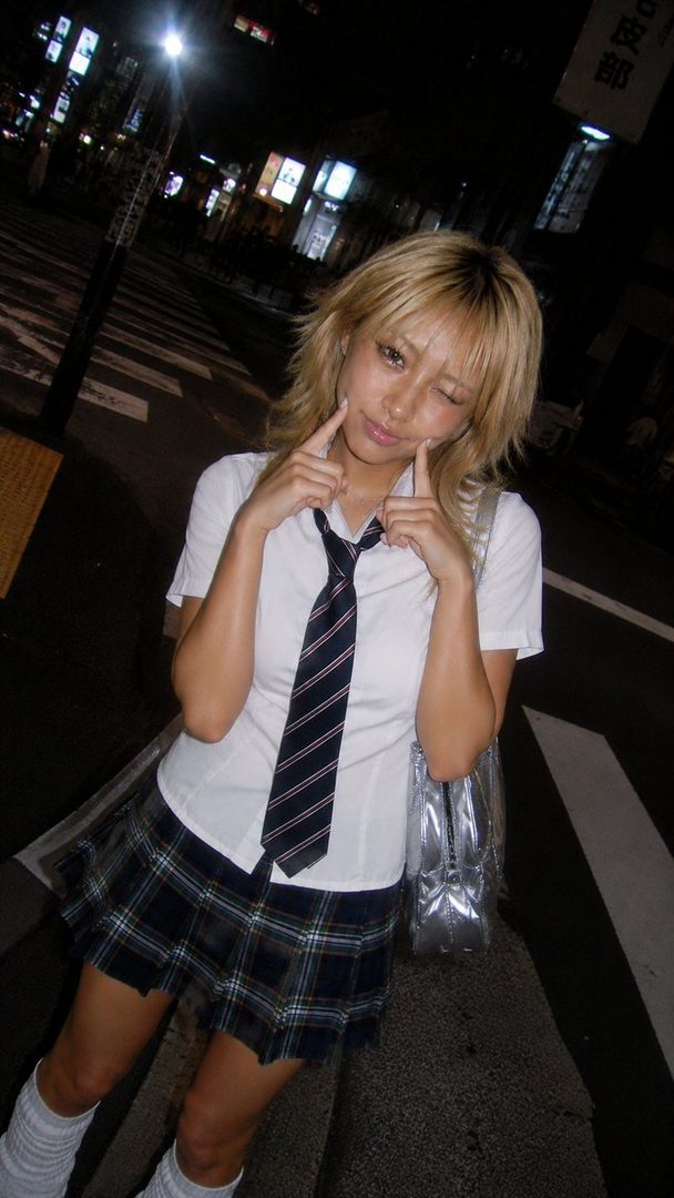 A young woman with blonde hair winks and poses with her fingers on her cheeks, wearing a white shirt, tie, and plaid skirt, standing on a city street at night.