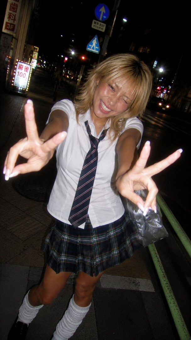 A young woman with blonde hair winks and makes a peace sign while wearing a school uniform at night on a city street.