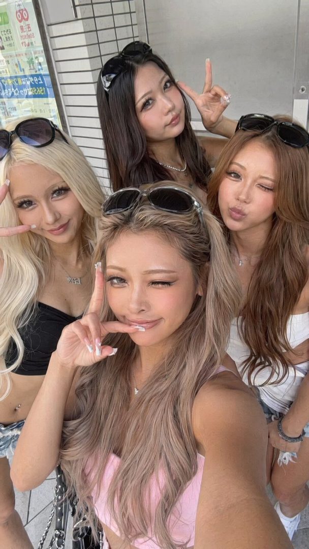 Four young women with long hair and sunglasses pose for a selfie outdoors, making peace signs.