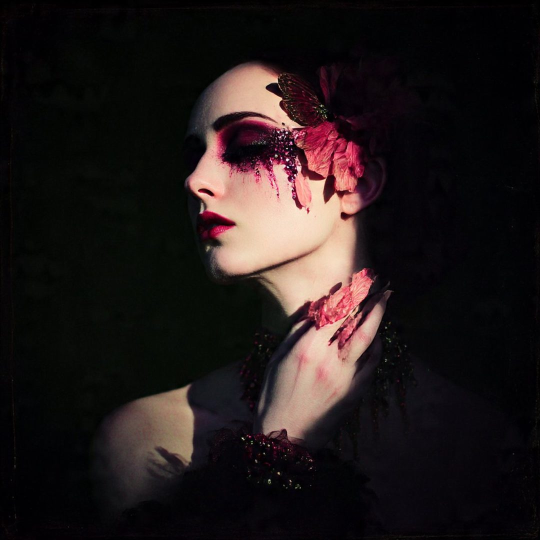 A woman with dramatic dark eye makeup and floral accessories poses elegantly.
