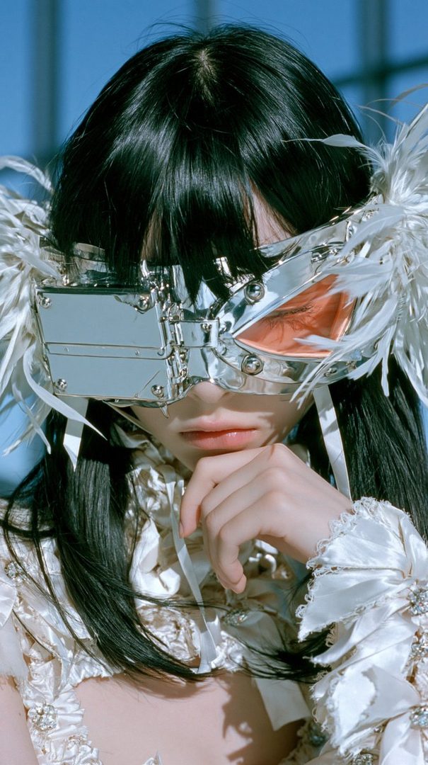 A person with long black hair wears a futuristic silver mask adorned with white feathers and a ruffled white outfit.