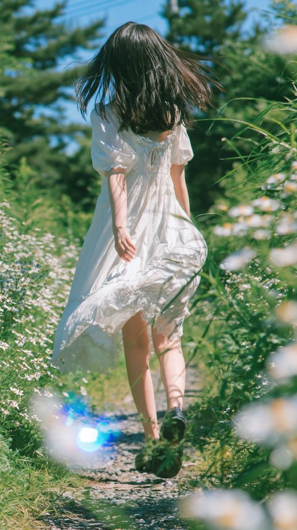 A young woman with long black hair, wearing a white dress, walks down a path lined with flowers and greenery.