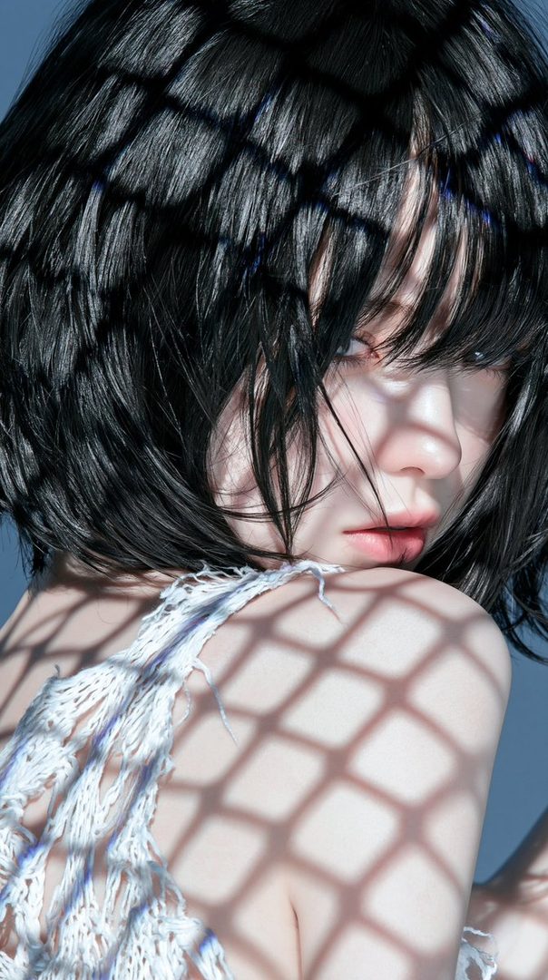 Close-up portrait of a young woman with short black hair, her face partially obscured by shadows, wearing a white top.
