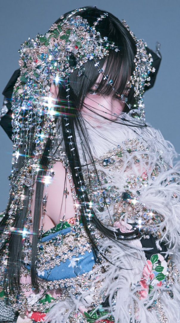 A woman with long black hair and bangs, adorned with an elaborate, sparkling headdress and ornate clothing, poses elegantly in a studio setting.