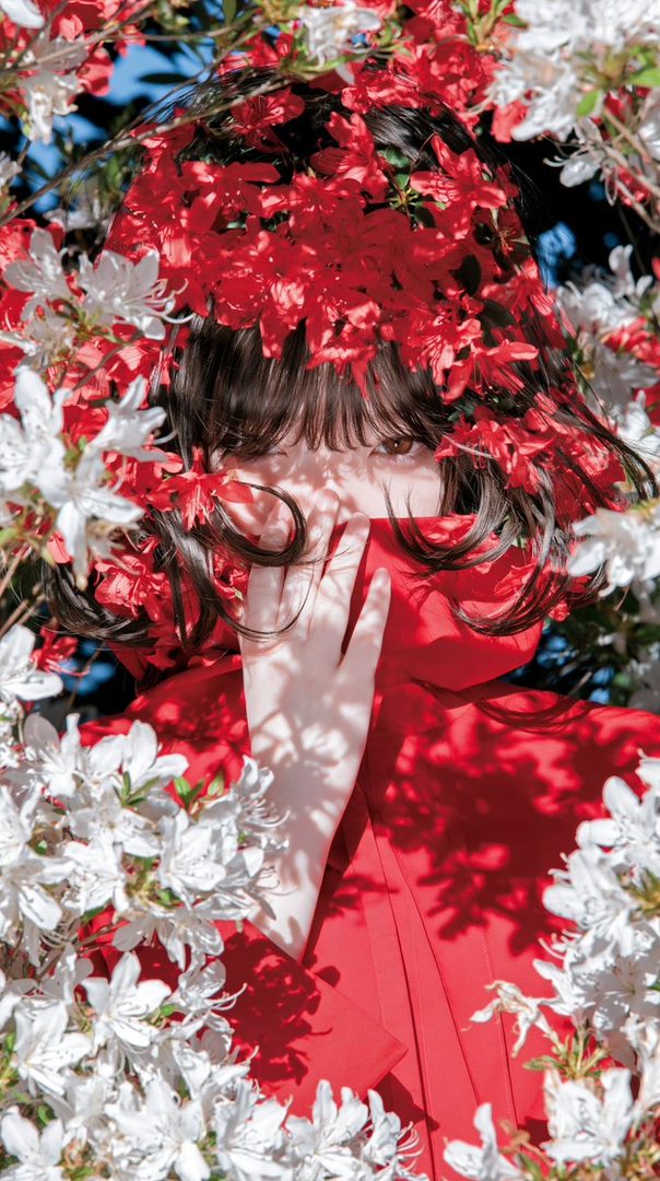 A portrait of a person with dark hair and bangs, partially obscured by red and white flowers, wearing a red dress.