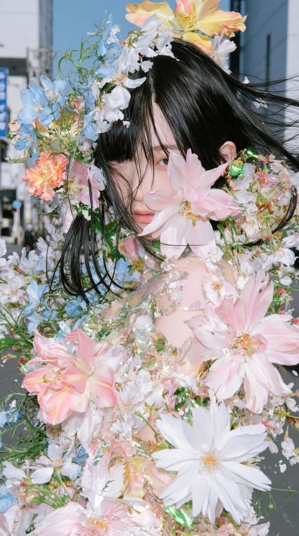 A portrait of a person adorned with a large floral arrangement, with dark hair and a delicate expression, set against an urban background.