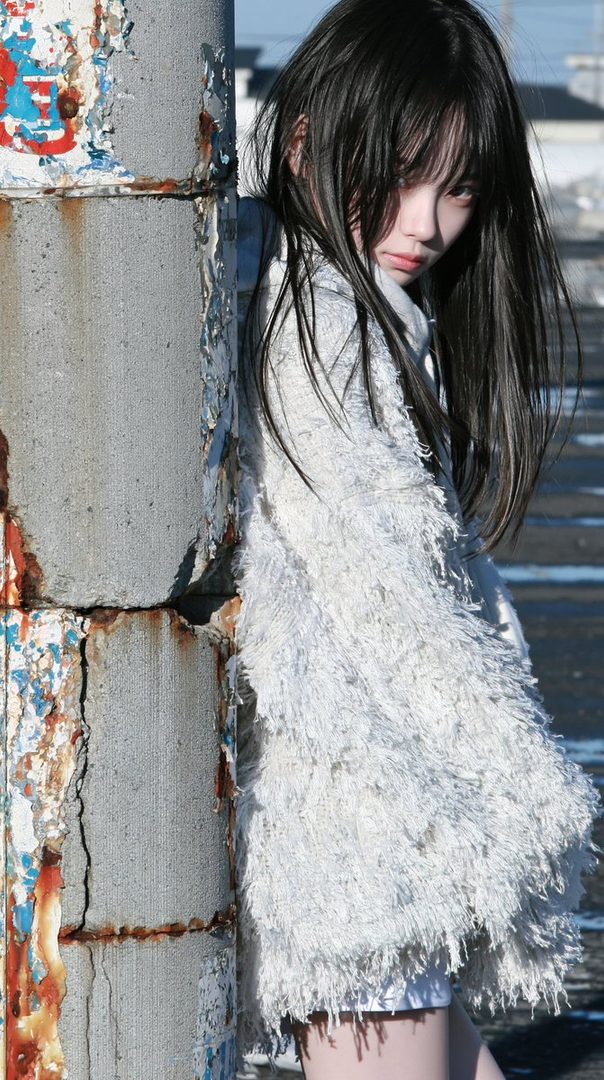A young woman with long black hair, wearing a fluffy white coat, leans against a weathered concrete pillar outdoors.