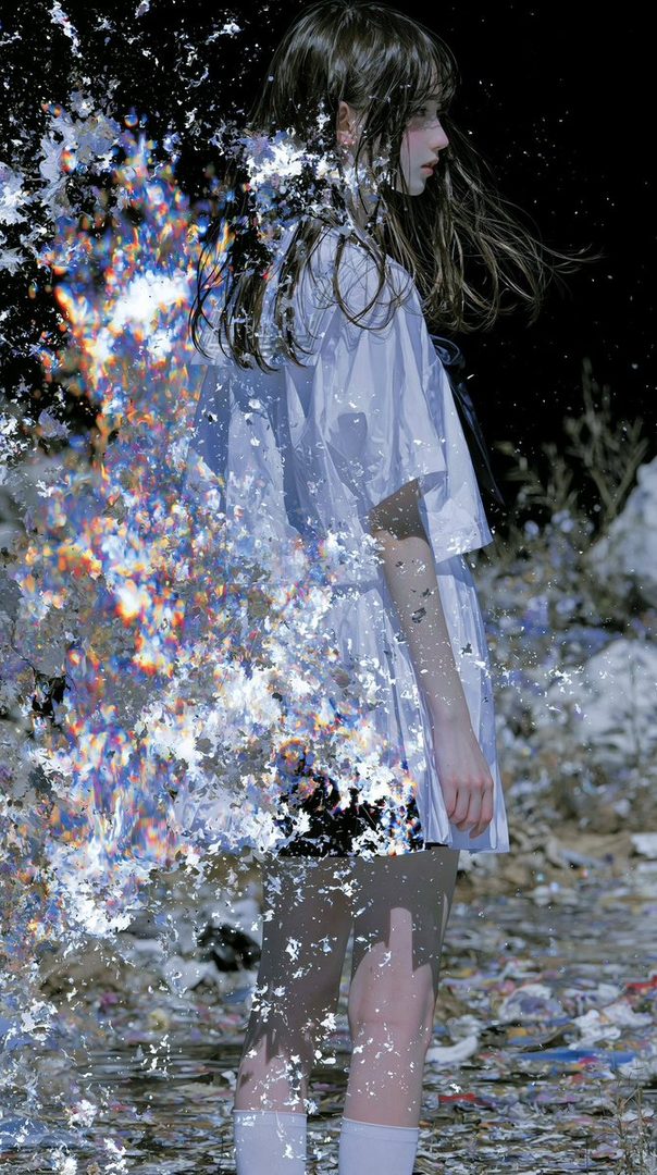 A young woman in a white school uniform stands outdoors at night, her body dissolving into colorful, fragmented light.