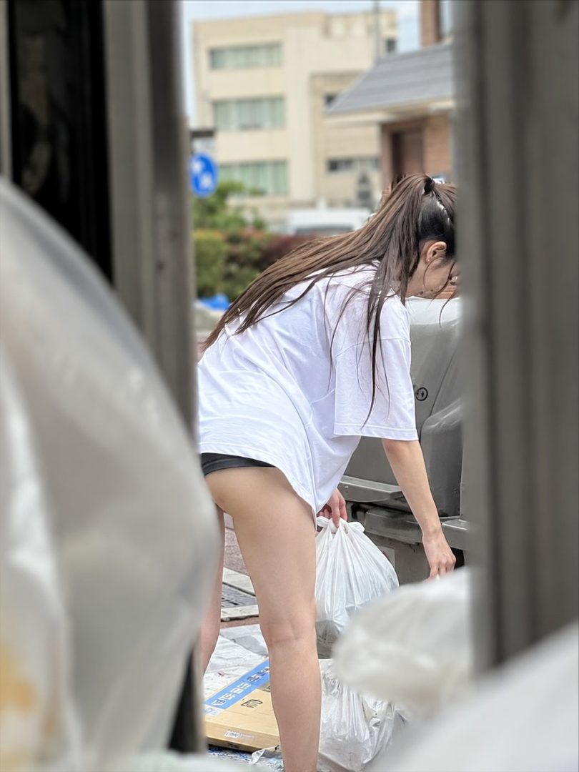 A young woman with long brown hair tied in a ponytail, wearing an oversized white t-shirt and black shorts, is seen from behind as she sorts through trash bags on a street.