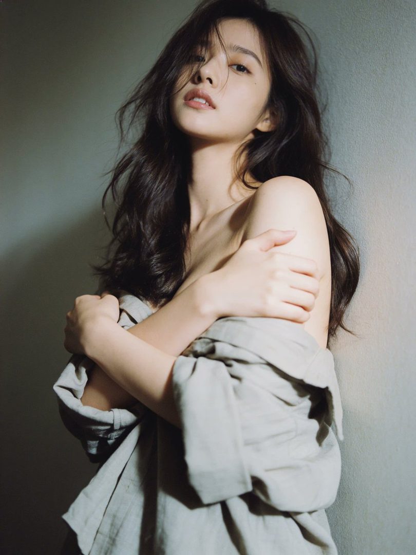 A young woman with long, dark hair is posing indoors, partially covered by a light-colored shirt, with a soft, dreamy expression.