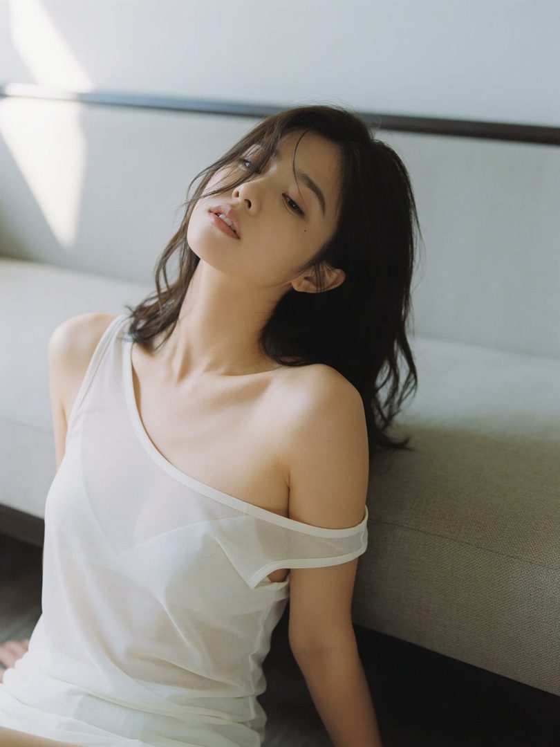 A young woman with long, dark hair poses indoors, wearing a white off-the-shoulder top, with soft morning light illuminating her face.