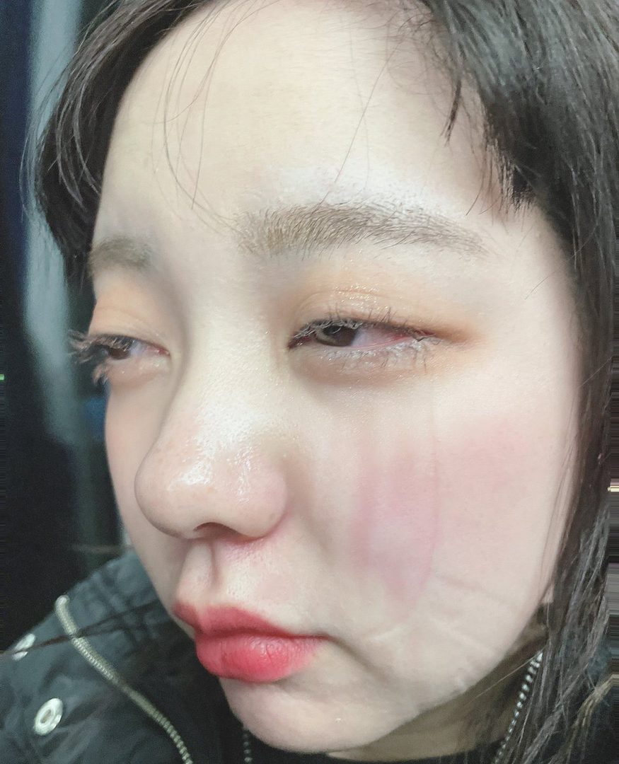 A close-up portrait of a young East Asian woman with short dark hair, looking down with a pensive expression, showing subtle makeup and a hint of red lipstick.