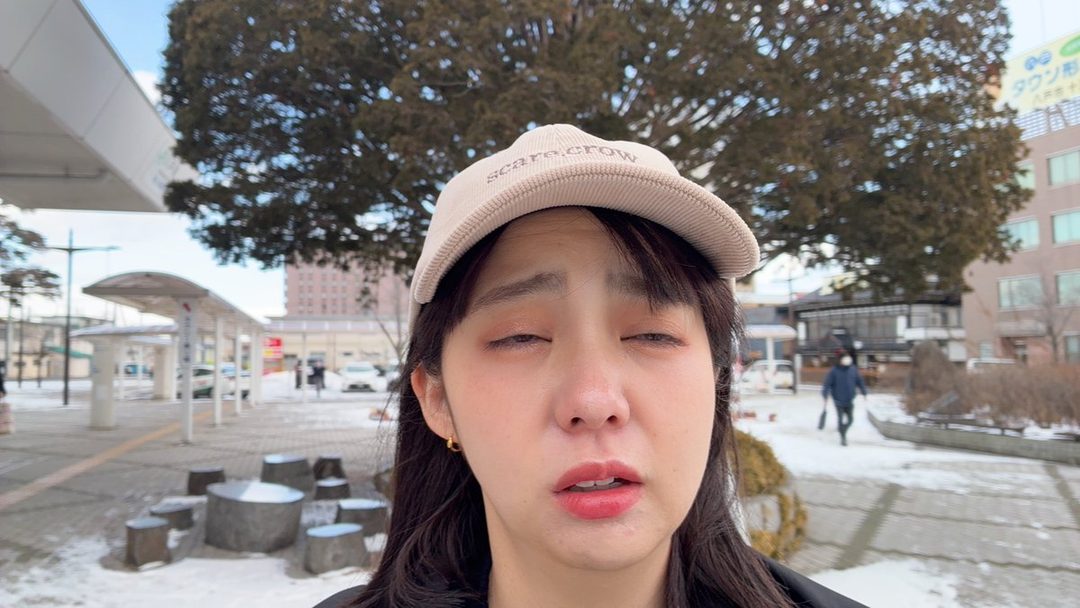 A young East Asian woman wearing a beige baseball cap with 'scare crow' embroidered on it, with her eyes closed and mouth slightly open, in an outdoor snowy urban setting.