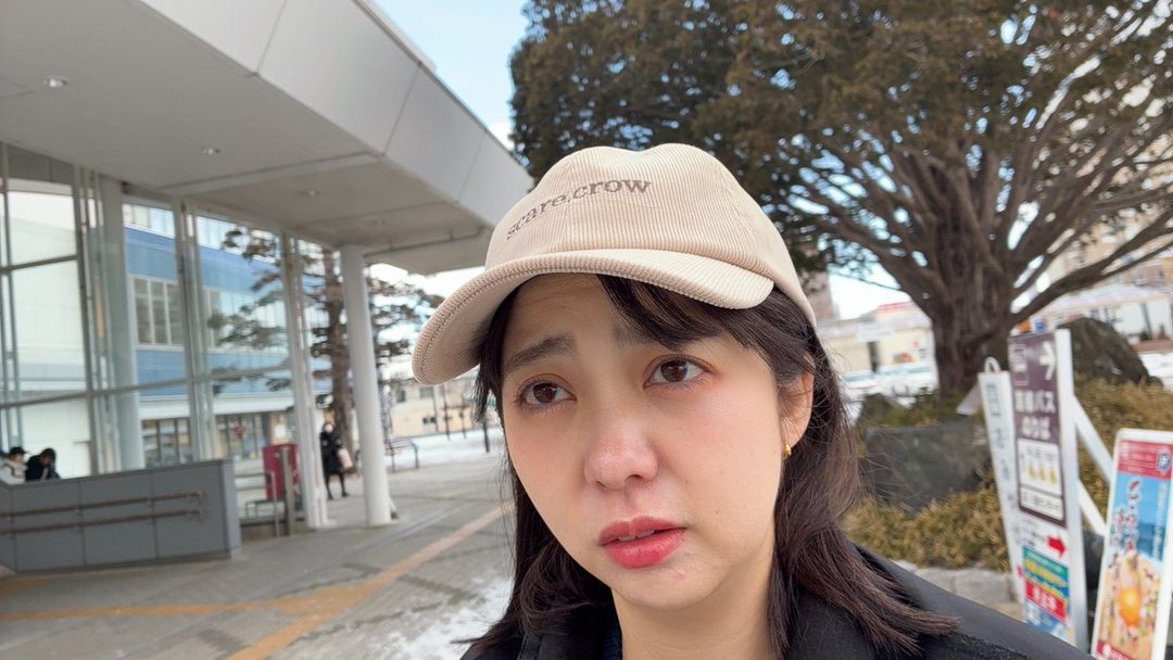 A young East Asian woman wearing a beige cap and a black jacket, looking slightly off-camera with a neutral expression, standing outdoors in front of a modern building.