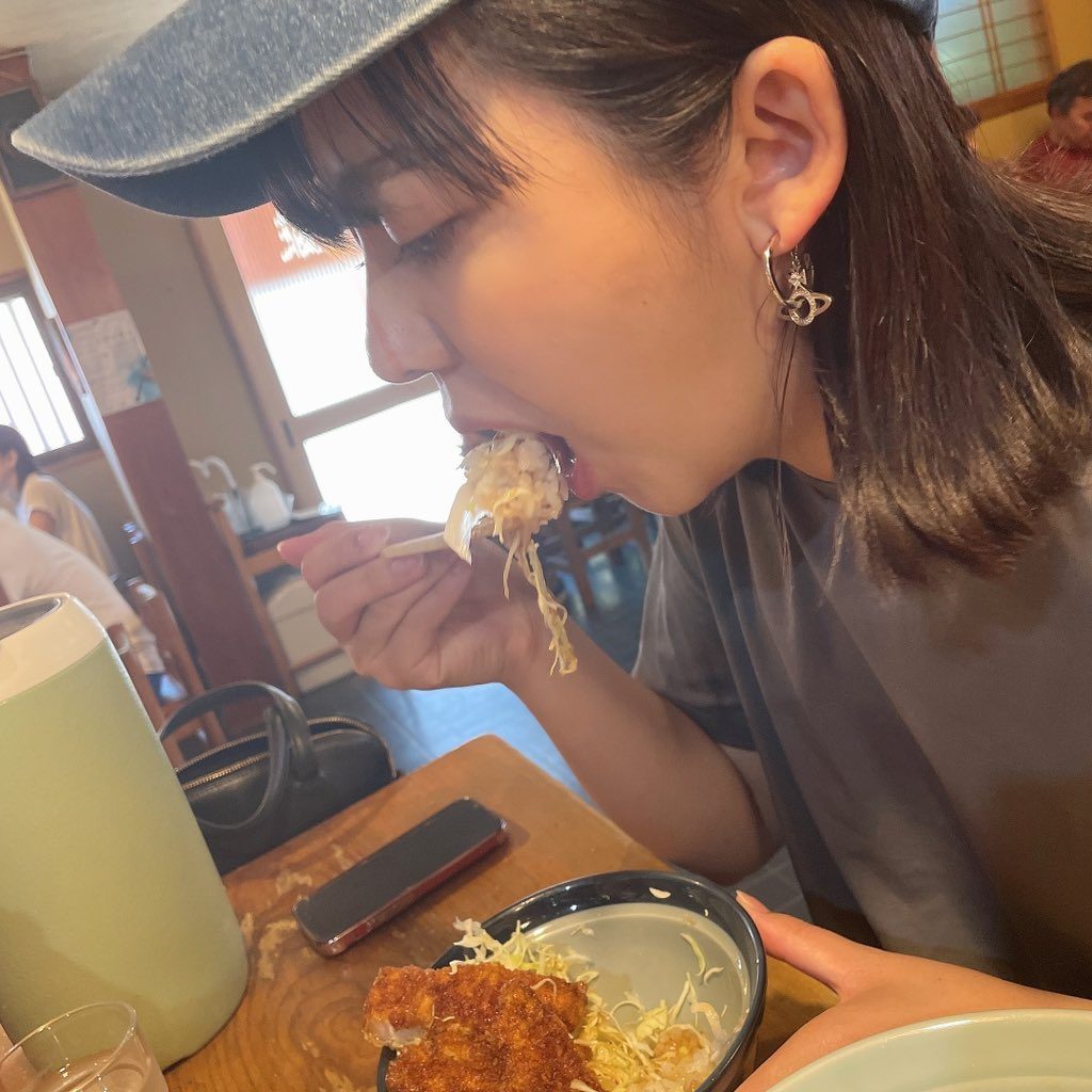 A young woman wearing a denim cap and a grey t-shirt eats food with chopsticks in a restaurant.