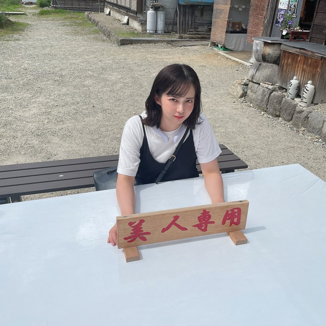 A young East Asian woman with short black hair sits at a table outdoors, wearing a white t-shirt and a black camisole, with a wooden sign in front of her.