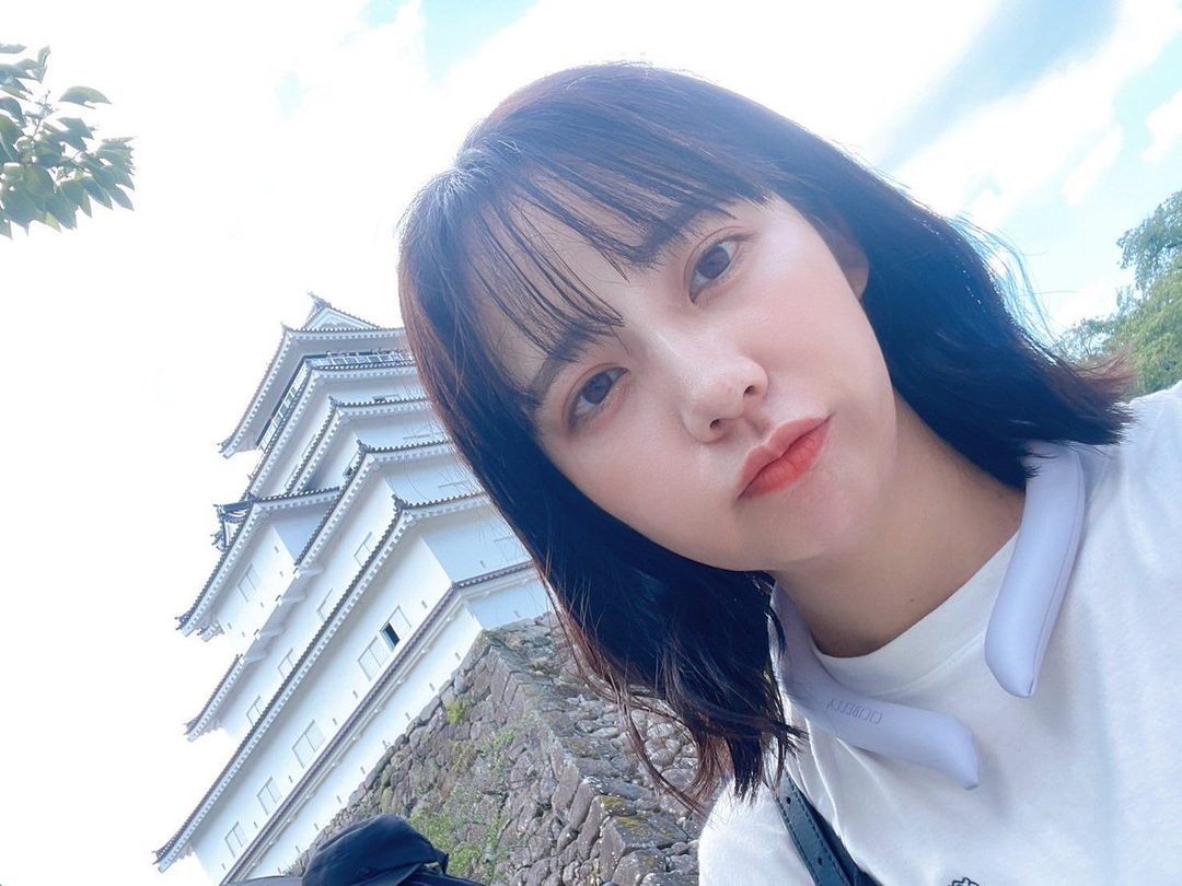 A young East Asian woman with short black hair takes a selfie in front of a traditional Japanese castle.