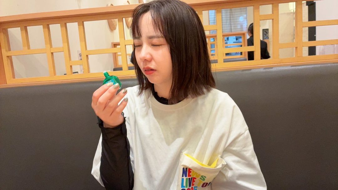 A young East Asian woman with short black hair, wearing a white t-shirt, holds a small green object in her hand and makes a funny face.