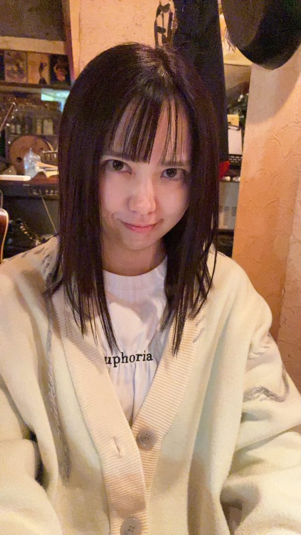 A young East Asian woman with shoulder-length dark hair and bangs smiles gently at the camera, wearing a white t-shirt with the word "uphoria" and a light-colored cardigan indoors.