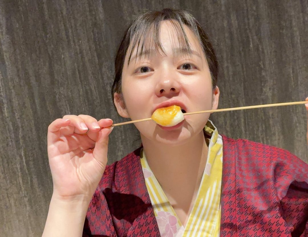 A young East Asian woman in a red and yellow yukata is eating a skewer with a round, white food item covered in a golden sauce.