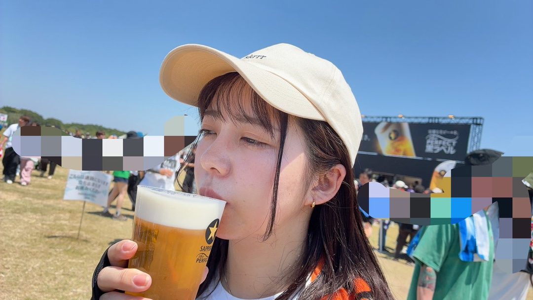 A young woman with long dark hair and bangs, wearing a beige baseball cap, drinks from a glass of beer outdoors.