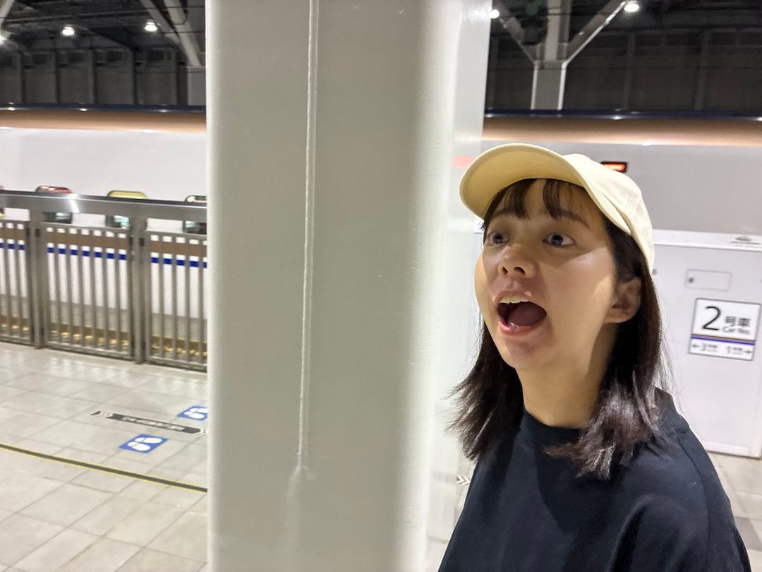 A young East Asian woman wearing a black t-shirt and a beige baseball cap, with her mouth open in surprise, stands in a train station.