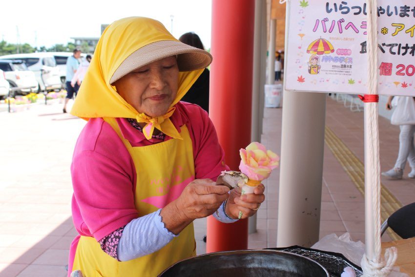 An elderly East Asian woman wearing a yellow sun hat and apron is carefully preparing a rose-shaped ice cream cone outdoors.