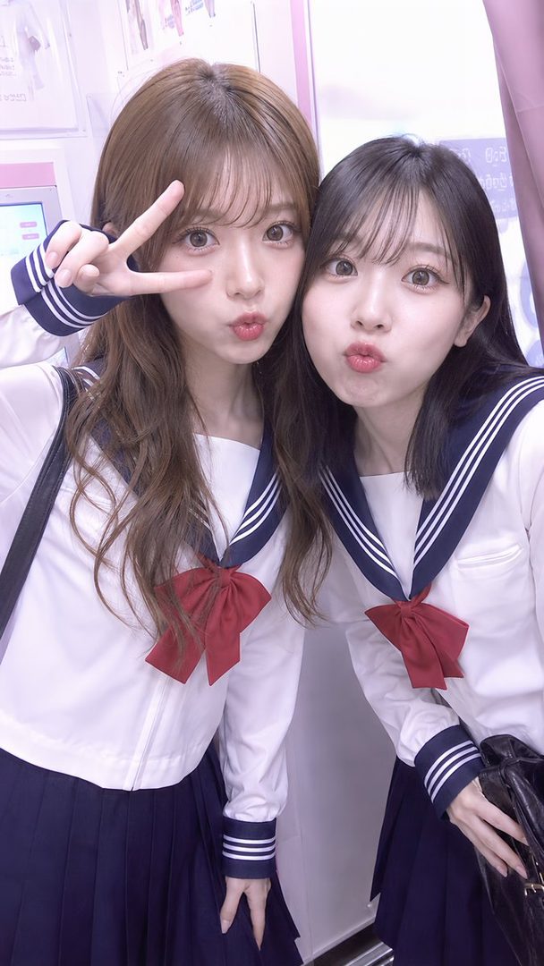 Two young women in Japanese school uniforms posing for a selfie with duck faces and peace signs.