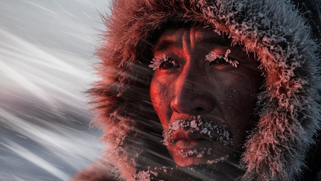 Extreme close-up of an East Asian person's face covered in frost and ice, wearing a fur-lined hood in a snowy blizzard.