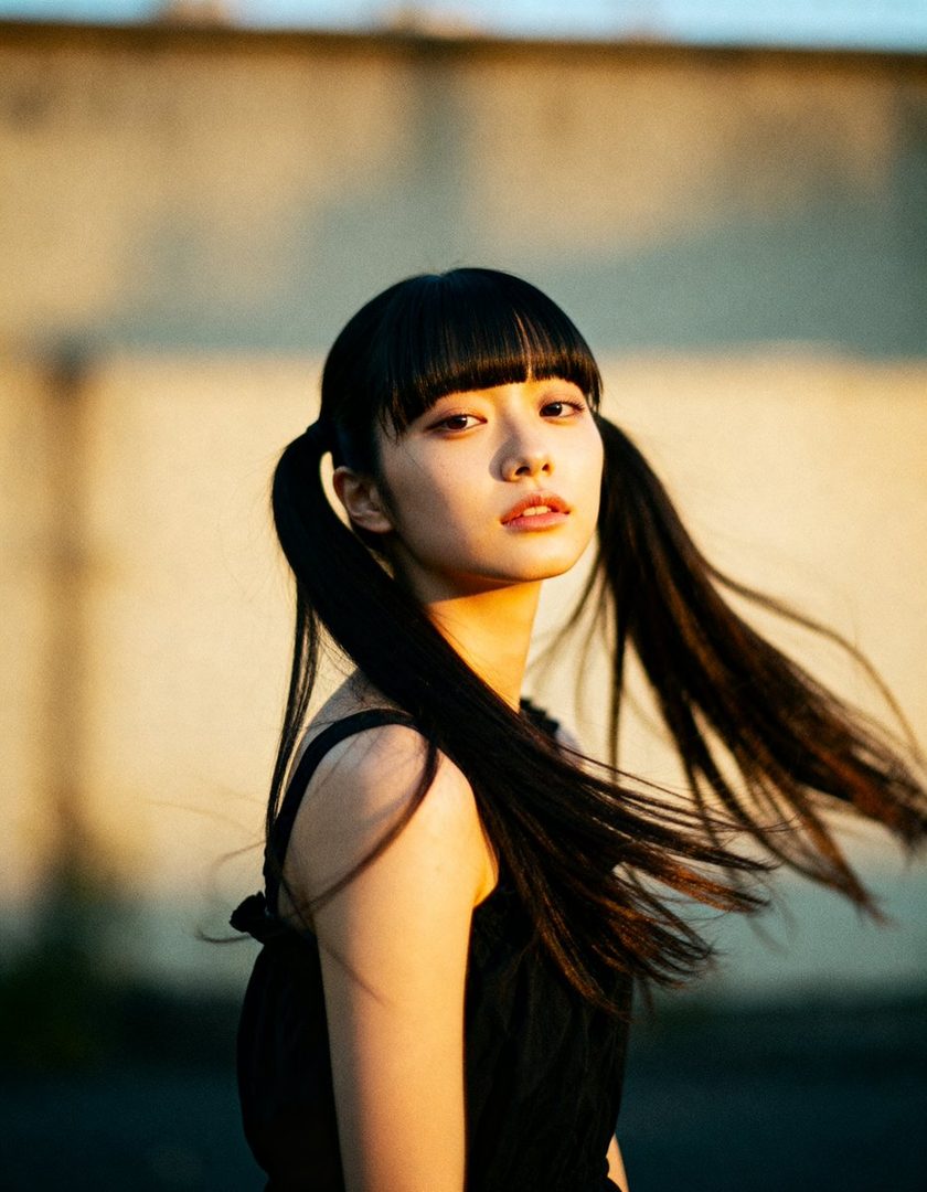 A young East Asian woman with black hair in twintails and bangs, wearing a black sleeveless top, poses outdoors in the golden hour light.