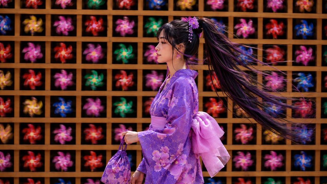 A Japanese woman with long black hair in a high ponytail, wearing a purple floral kimono and holding a matching drawstring bag, stands in profile against a wall of colorful pinwheels.