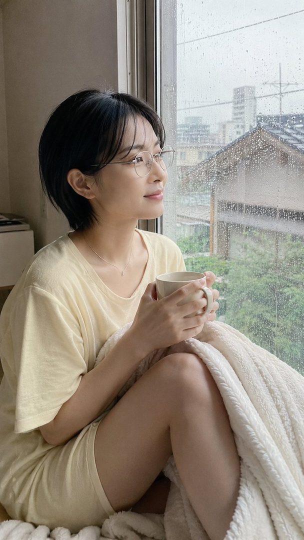A young woman with short black hair and glasses holds a mug of coffee while looking out a rainy window, wrapped in a blanket.