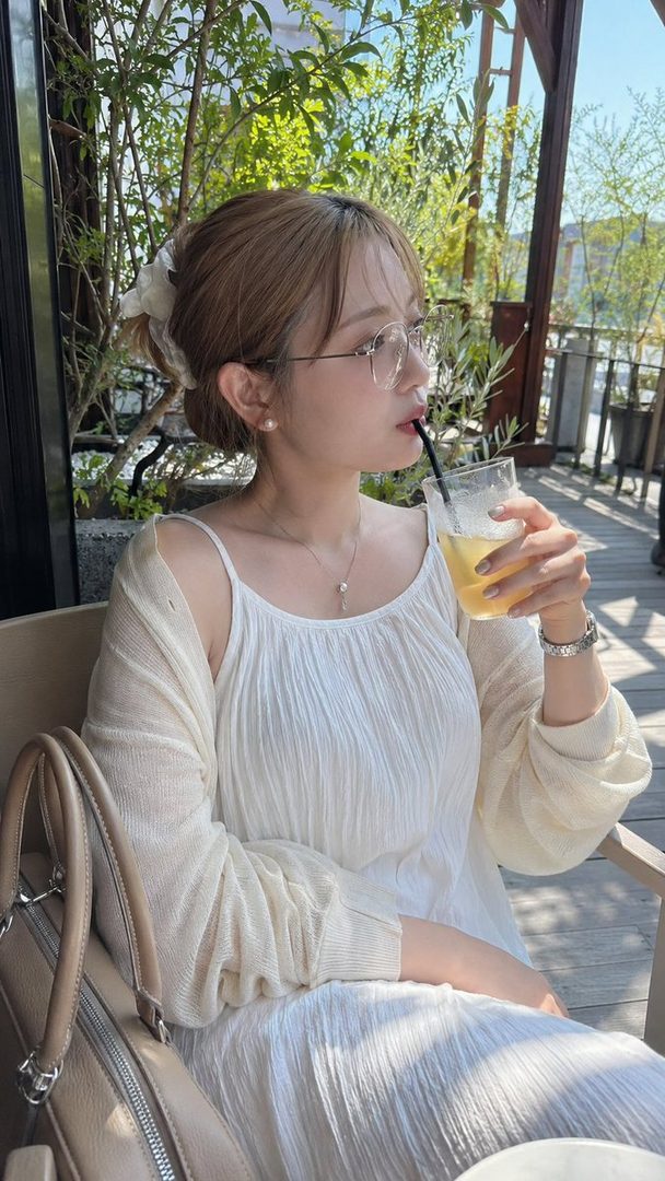 A young woman with brown hair tied up, wearing glasses and a white dress, drinks from a glass on a sunny terrace.