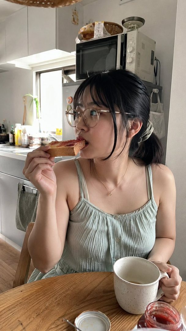 A young woman with black hair tied in a ponytail and wearing glasses, eating toast with jam and holding a mug, in a kitchen setting.