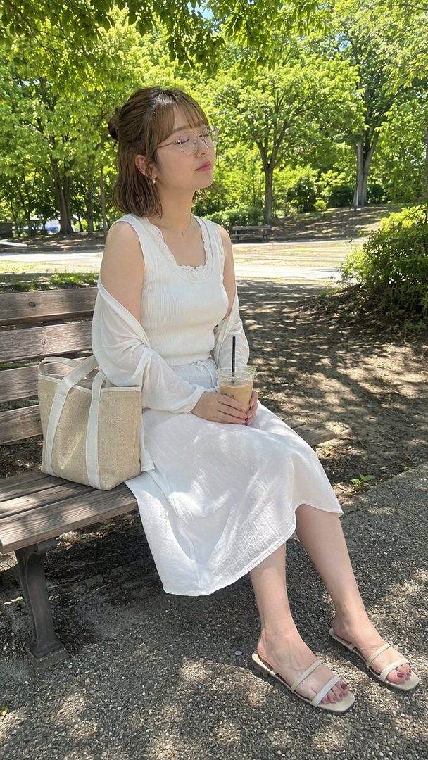 A young Japanese woman with short brown hair and glasses sits on a park bench, holding a cold drink and enjoying the shade.