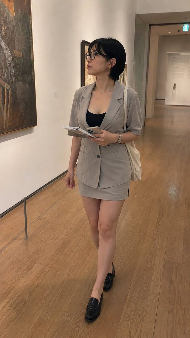 A woman with short black hair and glasses, wearing a light grey blazer and skirt, stands in an art museum looking at a painting.