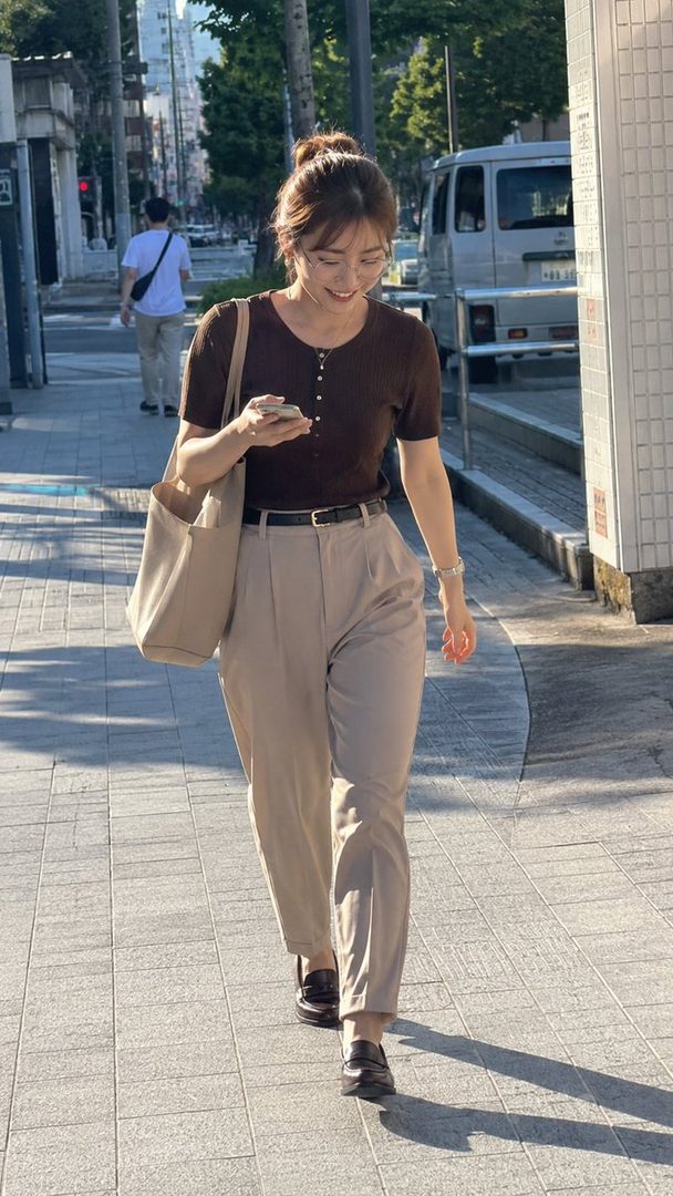 A young East Asian woman with her hair in a bun, wearing glasses, a brown short-sleeved shirt, and beige pants, walks down a city street while looking at her phone.