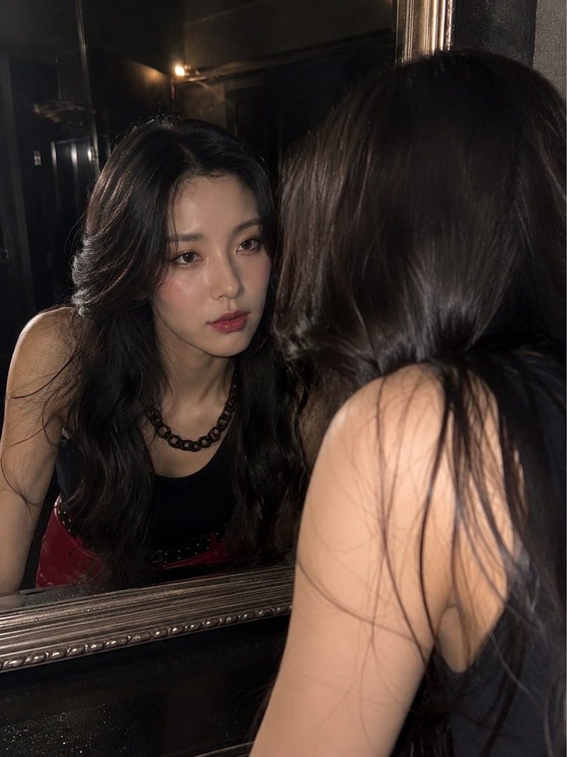 A young woman with long, dark, wavy hair looks into a mirror, wearing a black tank top and a chunky necklace.