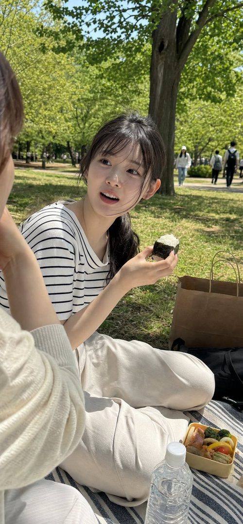 A young Japanese woman with black hair and bangs, wearing a striped t-shirt, sits on a blanket in a park, holding an onigiri and looking at someone off-camera.