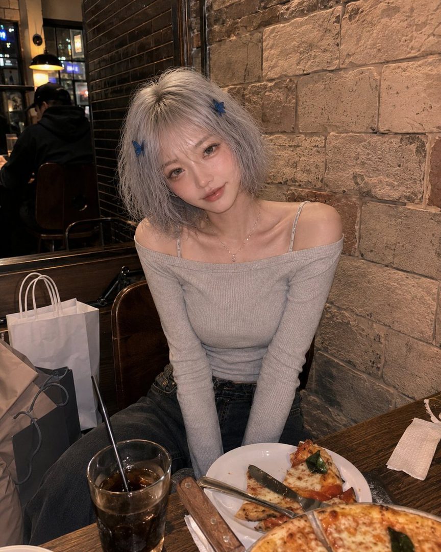 A young woman with short, silver hair and blue butterfly clips sits at a table with pizza and a drink, looking at the camera.