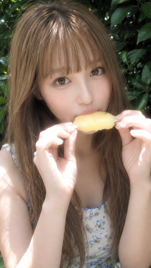A young woman with long brown hair and bangs is eating a piece of yellow fruit outdoors, wearing a white floral dress.