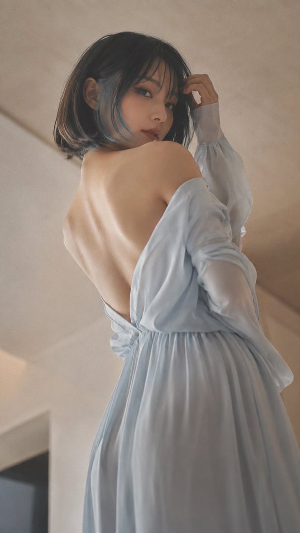 A woman with dark brown hair and blue highlights wears a light blue off-shoulder dress, looking back over her shoulder with her bare back exposed.