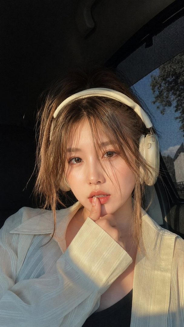 Close-up portrait of a young East Asian woman with brown hair and headphones, sitting in a car during golden hour, touching her lip.