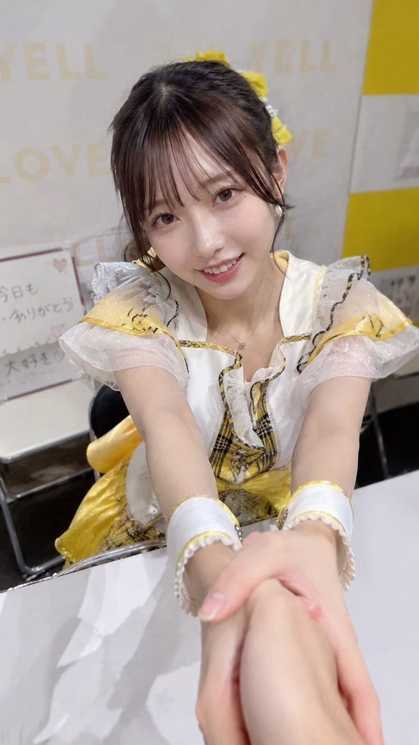 A young East Asian woman with brown hair tied up, wearing a yellow and white idol outfit, smiles at the camera while holding hands with someone.