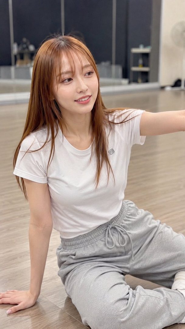 A young East Asian woman with long brown hair and bangs, wearing a white t-shirt and grey sweatpants, sits on a wooden floor in an indoor studio, smiling.